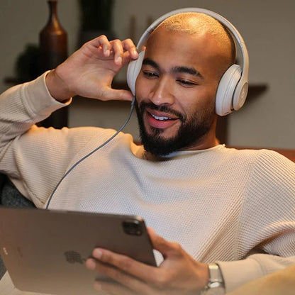 wearing Beats Studio Pro Kenya headphones in sand beige, connected via cable to an iPad in a relaxed indoor setting